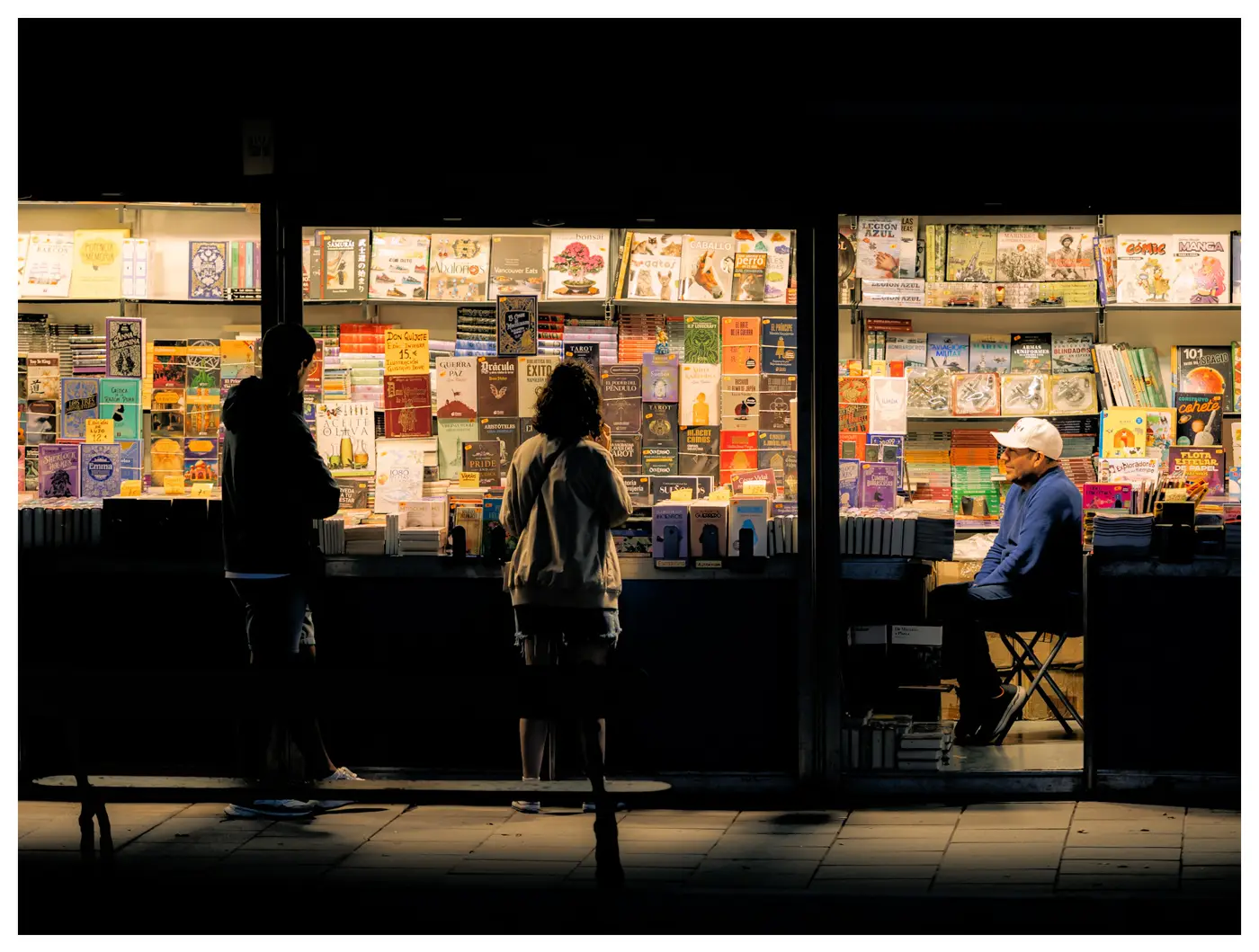 Dos personas miran libros en una caseta iluminada de la Feria del Libro de A Coruña, de noche, mientras el librero espera sentado en los Jardines de Méndez Núñez