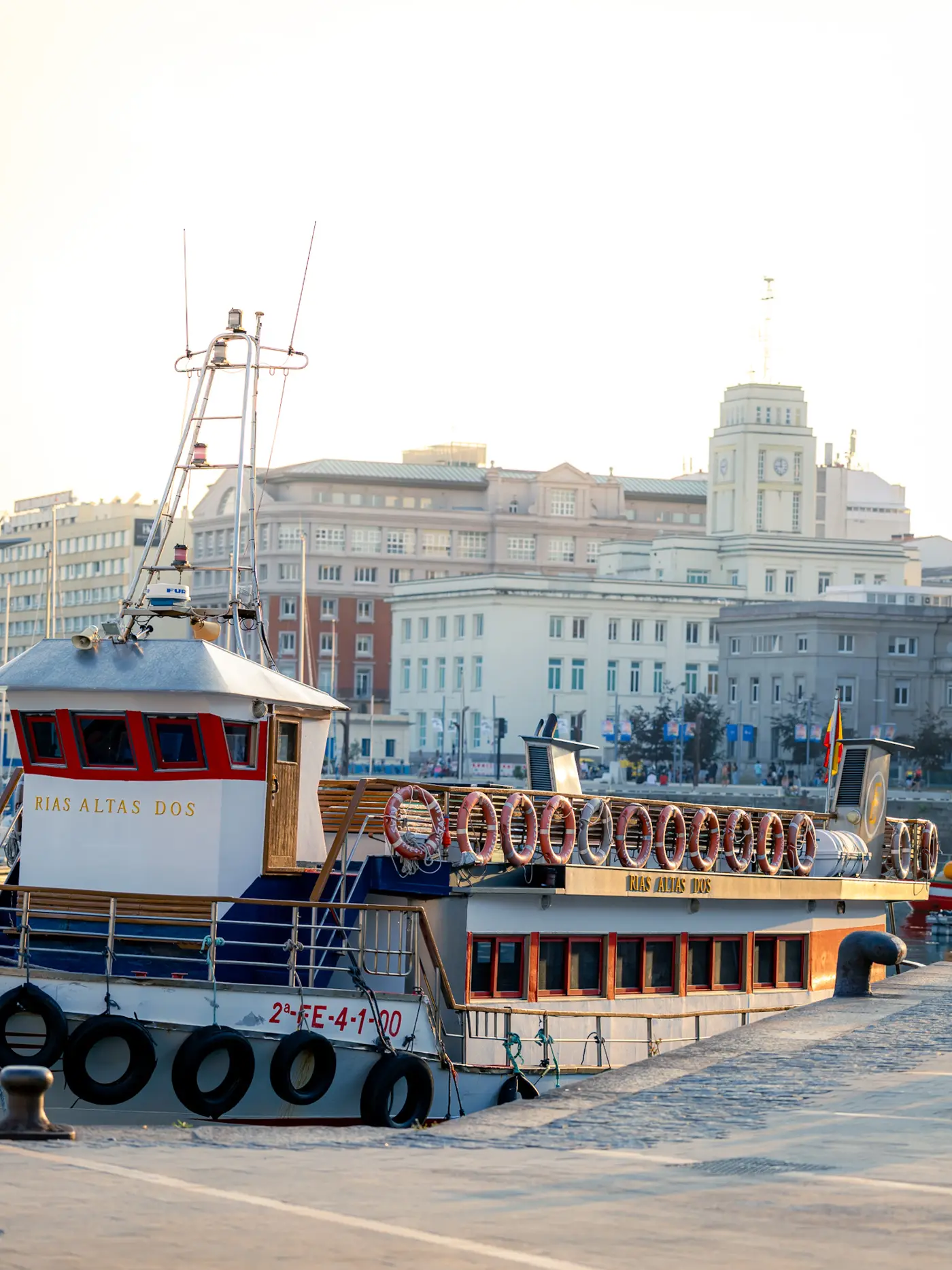 El barco de pasaje Rías Altas Dos amarrado en el puerto de A Coruña al atardecer, con sus salvavidas alineados en la cubierta y los edificios del paseo marítimo al fondo